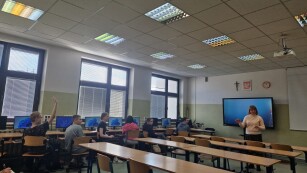 nauczyciel mówi do uczniów, którzy słuchają siedząc przy stanowiskach komputerowych a jeden uczeń zgłasza się do odpowiedzi
