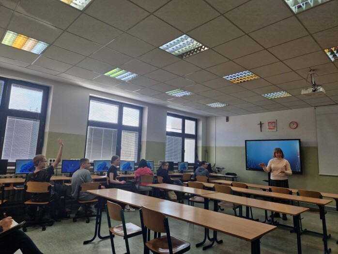 nauczyciel mówi do uczniów, którzy słuchają siedząc przy stanowiskach komputerowych a jeden uczeń zgłasza się do odpowiedzi