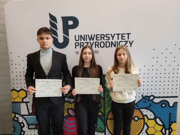 Trzej uczniowie na tle banneru Uniwersytetu Przyrodniczego w Lublinie z certyfikatami uczestnictwa