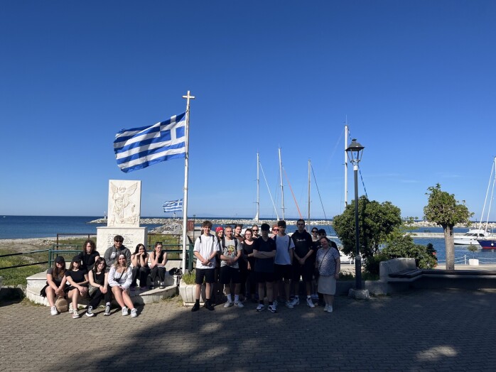 Grupa uczniów stoi na tle morza i flagi greckiej